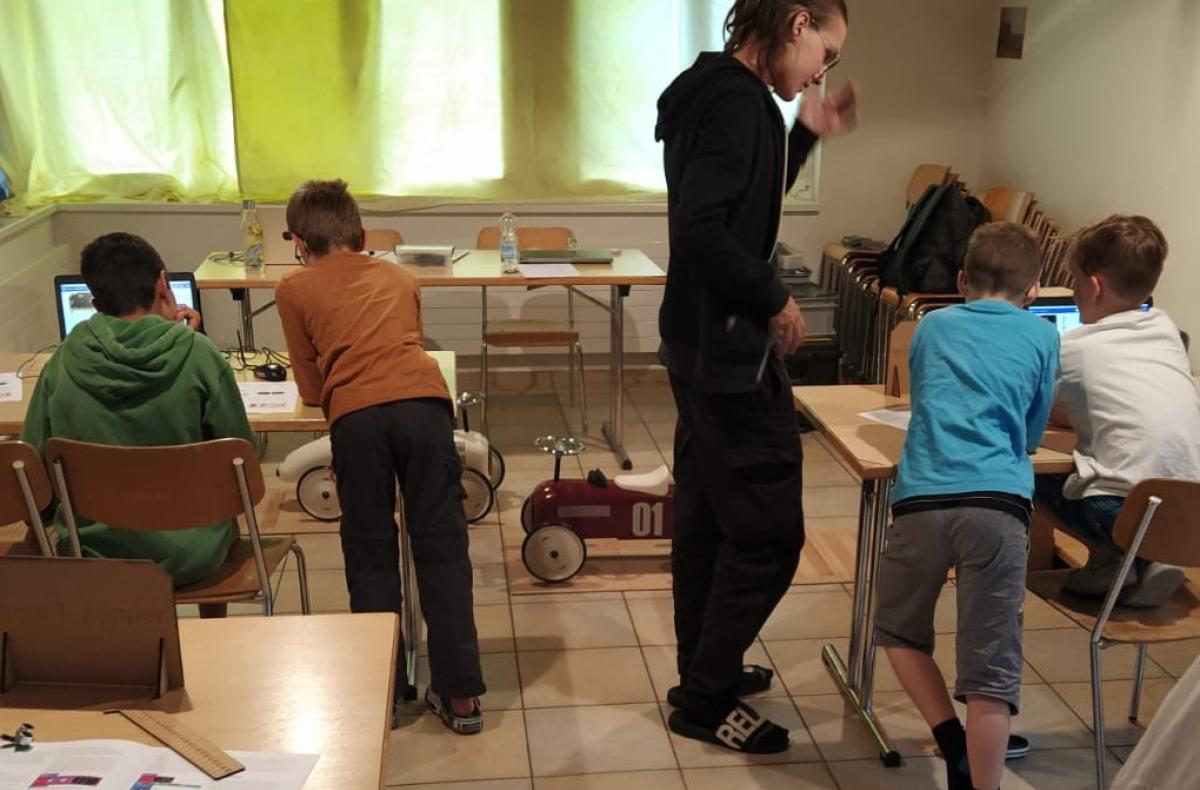 A coach works with a group of children at laptops to control model cars.