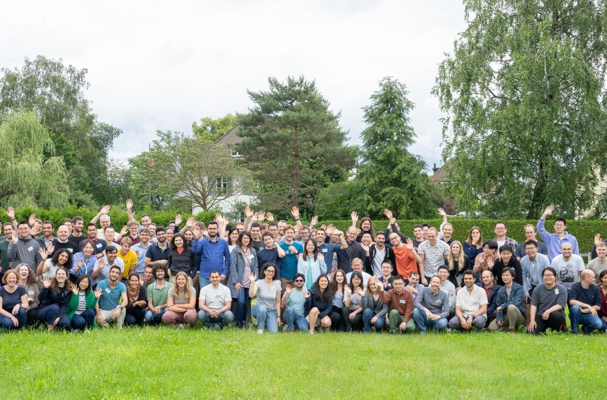 A large group of people pose outside, smiling and waving