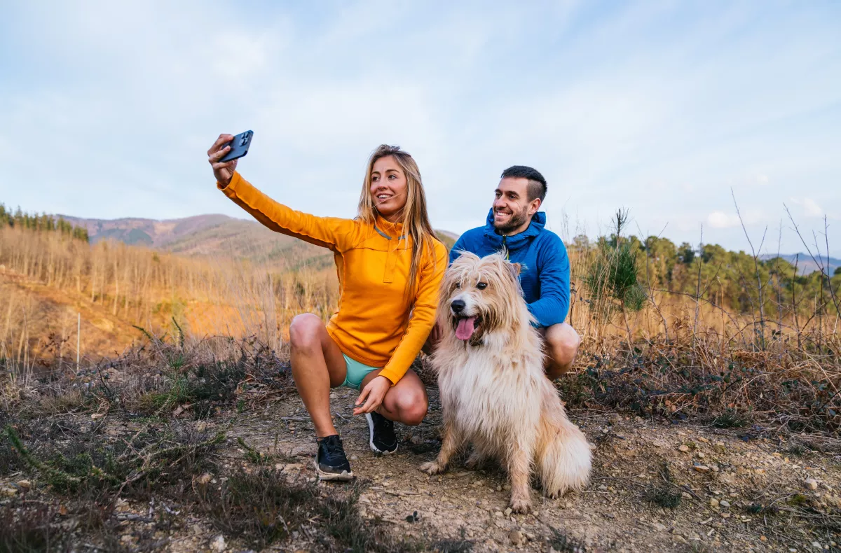 two people in a field taking a selfie of them and their dog using a mobile phone
