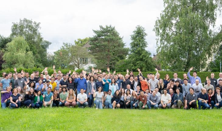 A large group of people pose outside, smiling and waving 