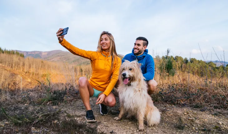 two people in a field taking a selfie of them and their dog using a mobile phone
